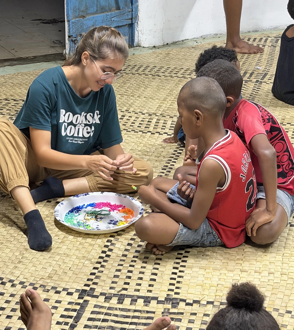 Rebecca sitting with children on a woven mat, making bracelets beside a tray of colorful beads.